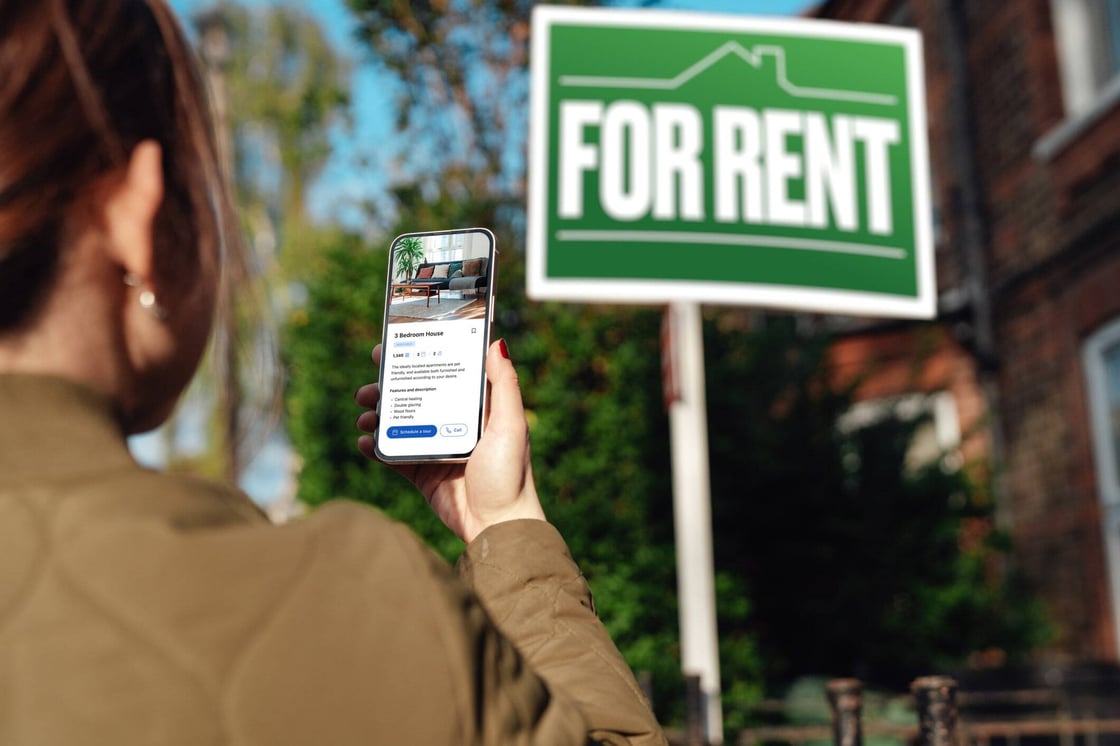 A woman looking at a rental listing on her phone with a “for rent” sign in the background.