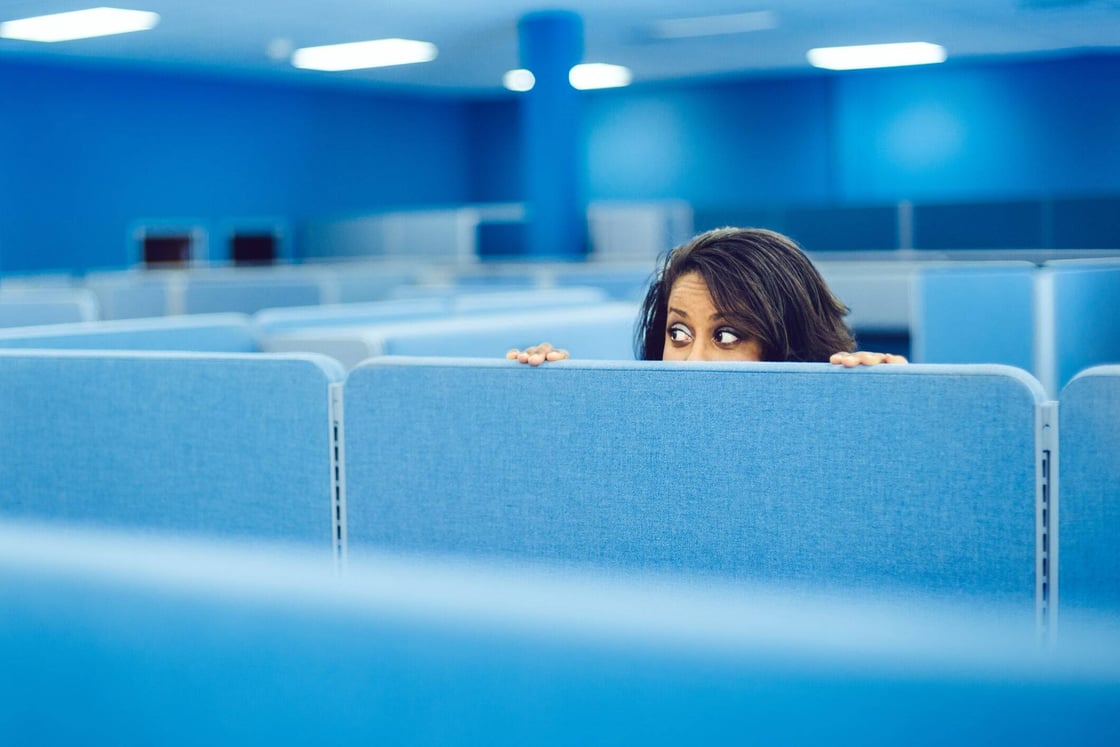 A woman peeking out from behind a blue office cubicle.