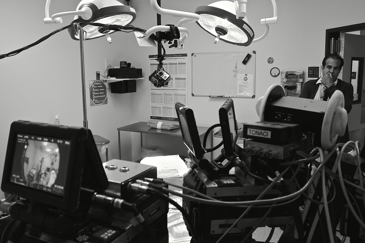 Operating room in black and white: Ted Gagliano surveys, with two illuminated monitors and Tran's cameras including a tripod-mounted camera; surgeon reaching for tool, instruments ready; medical equipment and surgical light overhead.