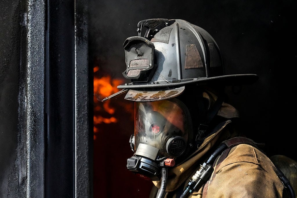 Close-up of a firefighter in a helmet and breathing mask, looking right, with visible flames in the dark background.