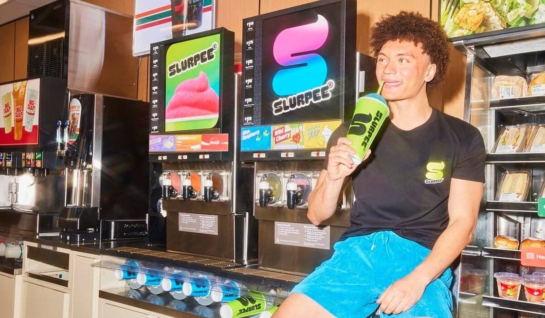 A young man drinks a Slurpee leaning next to a Slurpee machine.