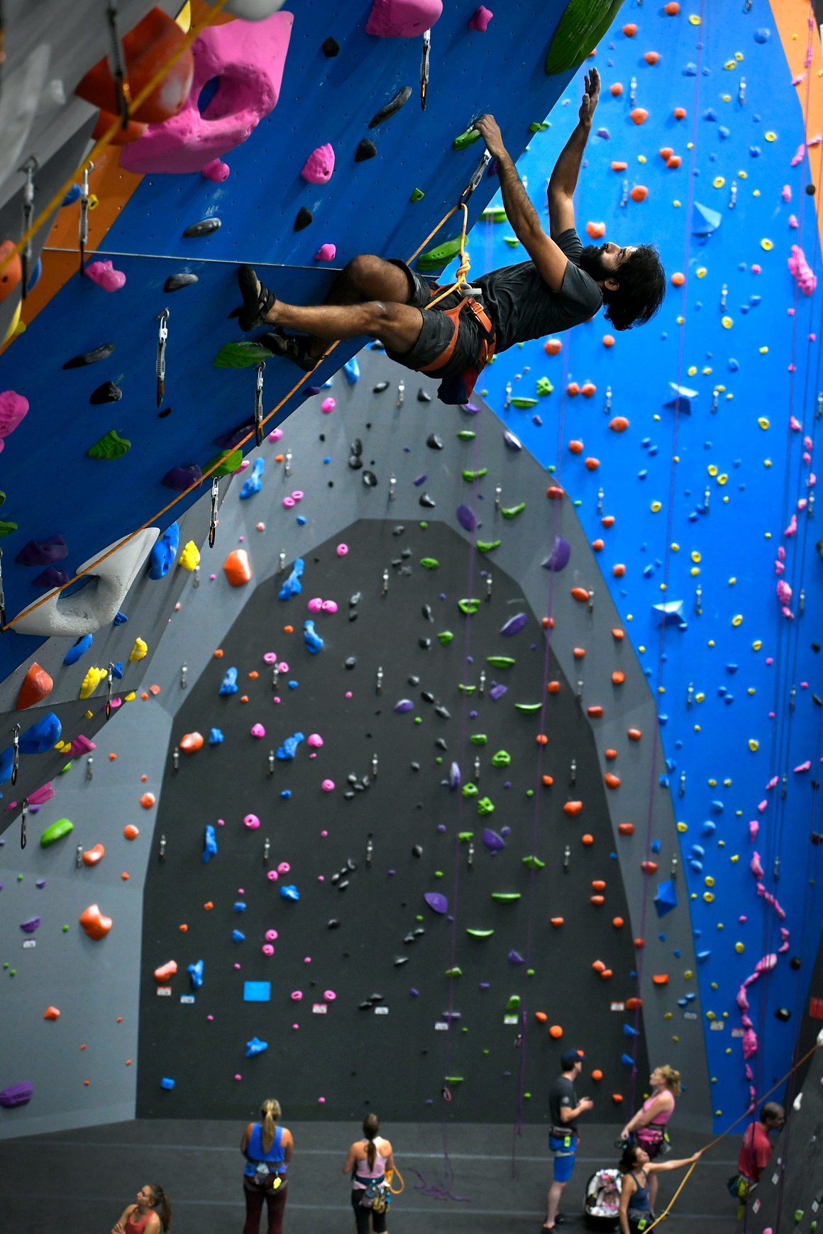 A man hangs mid-air while lead climbing on a tall, colorful indoor rock wall. Several people are visible below.