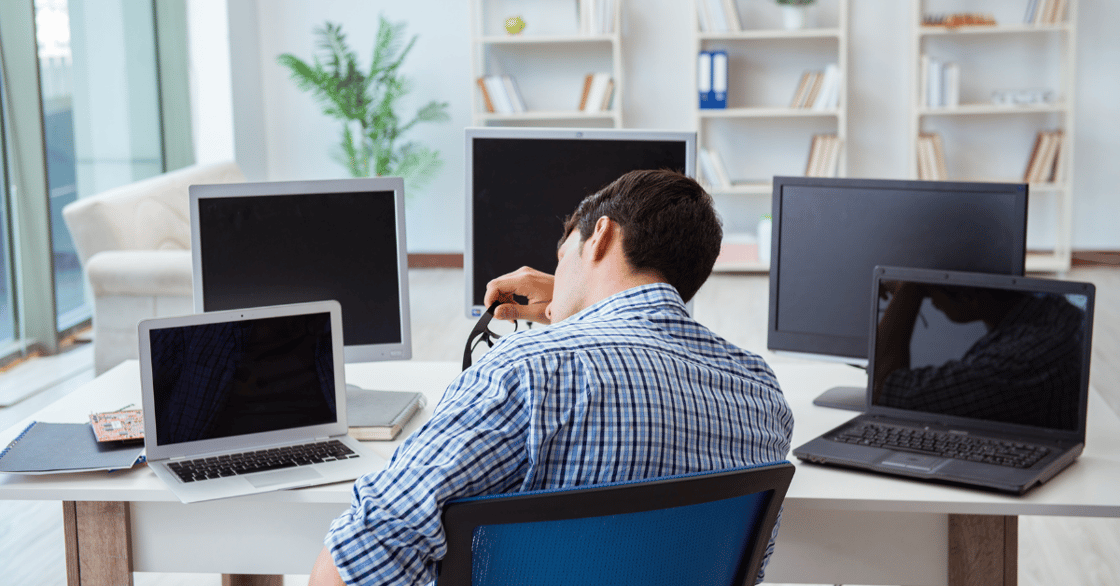 A man rubs his face, looking exhausted, as he sits at a desk in front of several computers.
